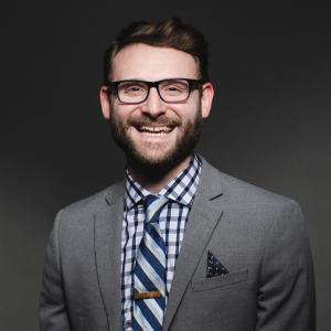 Doug Kowalewski wearing a plaid shirt, striped tie, and grey suit jacket smiling for his headshot.