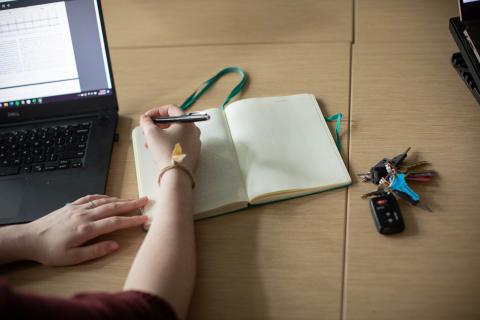 An image of a hand writing in a notebook with a laptop open.
