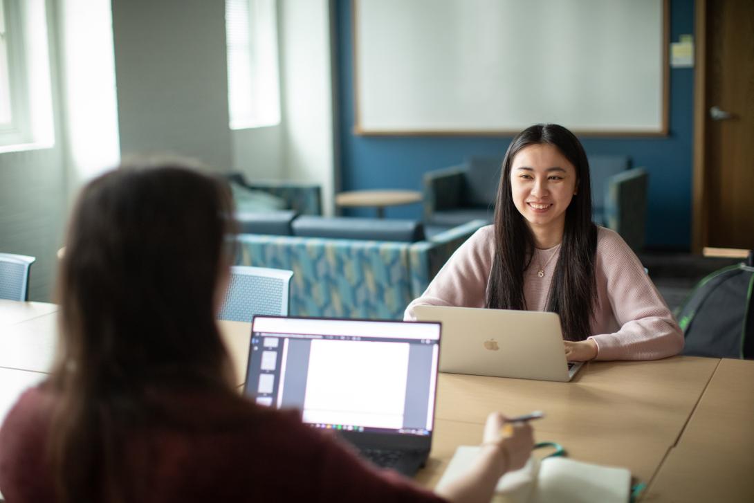 Two students sitting at a conference table talking with their laptops open.